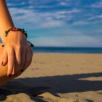 Woman sitting on the beach with closeup of hand with bracelet and nail polish and knee with shadow, blue sky and sea in the background summertime