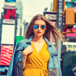 Young Eastern European Woman traveling in New York, with long brown hair, wearing yellow dress, blue Denim jacket draped over shoulder, blue sunglasses, standing on street in Times Square of Manhattan